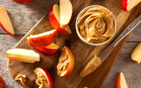 Saftige Apfelspalten mit cremiger Erdnussbutter auf einem Holzbrett. Ein Messer mit Erdnussbutter liegt daneben.