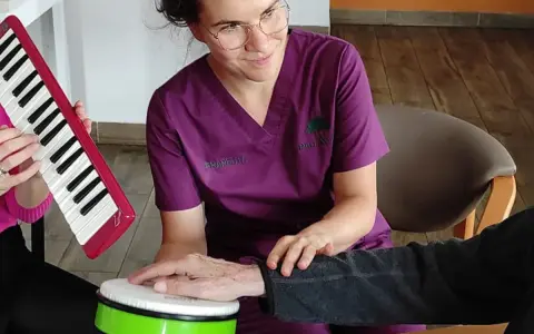 Terapia dźwiękiem i rytmem: muzykoterapia dla seniorów. Terapeuta wspiera dłoń pacjenta na bębenku, podczas gdy inny uczestnik gra na keyboardzie.
