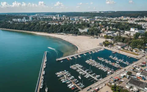 Widok z lotu ptaka na plażę miejską w Gdyni, z zatoką pełną jachtów i łodzi, oraz miastem w tle.