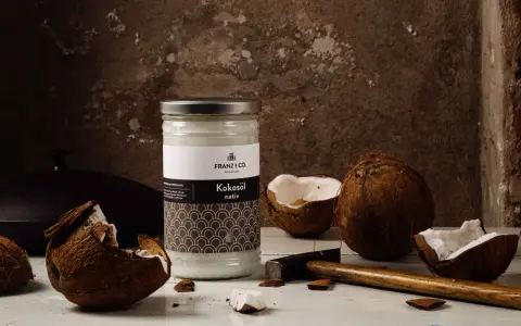 Ein Glas Kokosöl nativ steht neben zerbrochenen Kokosnüssen und einem Hammer. So wird aus der Frucht das wertvolle Öl gewonnen.