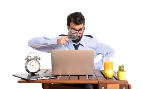 Mężczyzna z lupą bada laptopa, szukając odpowiedzi, czy skstore jest legit. Na biurku budzik, jabłko i sok.