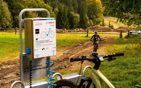 Stacje ładowania rowerów elektrycznych w Piwnicznej-Zdroju. W tle rowerzystka na szlaku.