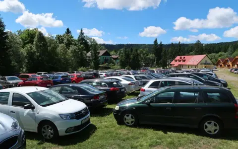 Tłum samochodów na parkingu przy Karłowie Szczeliniec. Słoneczny dzień, chmury na niebie, otoczenie lasu i budynków.
