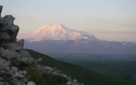 Elbrus, najwyższy szczyt Rosji, lśni w blasku zachodzącego słońca. W oddali widać zielone wzgórza i skaliste zbocze.