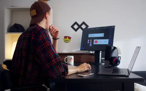 Młody mężczyzna w czapce i flanelowej koszuli pracuje przy komputerze. To typowy freelancer, który tworzy swoje projekty z domu.