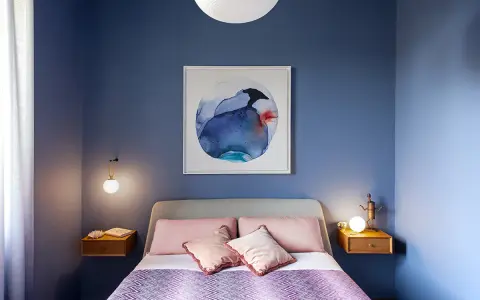 Camera da letto con pareti blu intenso e un quadro astratto. Cuscini rosa e coperta viola creano colori rilassanti.