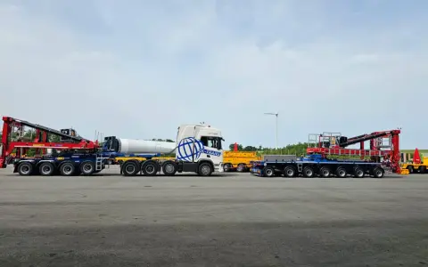 Ciężarówka z naczepą tandem przewozi element turbiny wiatrowej. Obok stoi inna naczepa z dźwigiem.