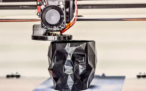 A 3D printer nozzle extrudes filament to create a faceted black skull planter, showcasing innovative 3D printing examples.