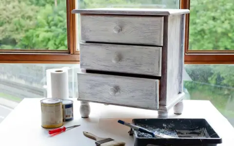 Mueble de tres cajones con acabado desgastado, listo para un proyecto de como pintar muebles. Pinceles, rodillo y latas de pintura a su lado.