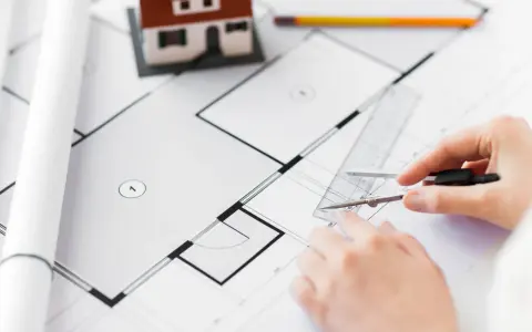 Rysowanie planu domu, przygotowanie do uzyskania warunków zabudowy. W tle makieta domu i zwinięte projekty.