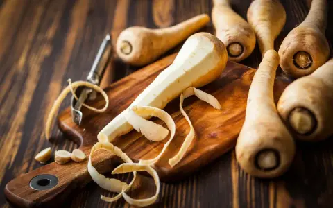 La pastinaca pianta, parzialmente pelata su un tagliere, circondata da altre radici e un pelapatate.