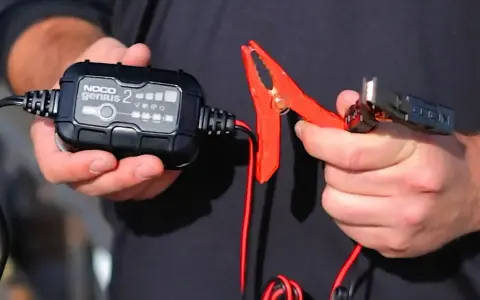 A person holds a NOCO Genius 2 trickle charger and its red clamp. This charger is designed to prevent damage, so no, a trickle charger can't ruin a battery when used correctly.