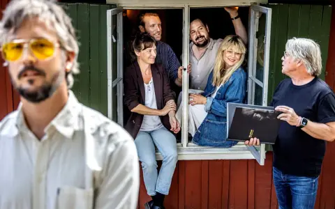 En man med gula glasögon och en grupp människor i ett fönster. En man håller en bok med texten "Allan Edwall barn".