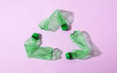 Trzy zielone, zgniecione butelki tworzą symbol recyklingu na różowym tle.
