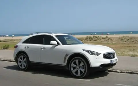 Białe infiniti auto zaparkowane przy plaży, z widokiem na morze i ludzi odpoczywających na piasku.