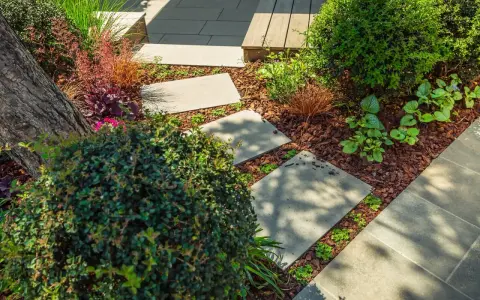 Schritt für Schritt: Gehwegplatten verlegen in einem üppigen Garten mit Rindenmulch und verschiedenen Pflanzen.