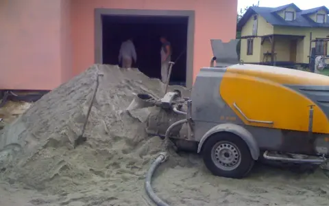 Miksokret przygotowuje zaprawę na budowie. Obok wielka kupa piasku, a w tle budynek i pracownicy.
