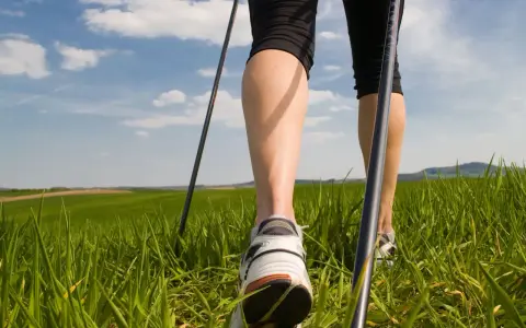 Gambe che praticano nordic walking tecnica su un prato verde sotto un cielo azzurro con nuvole bianche.