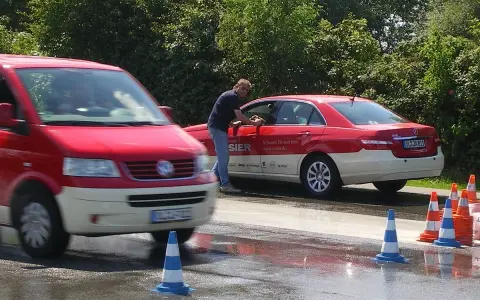 Mann reinigt Taxi. Ein roter Transporter fährt vorbei. Verkehrskegel markieren den Bereich. Möglicherweise eine Schulung der Berufsgenossenschaft Taxi.