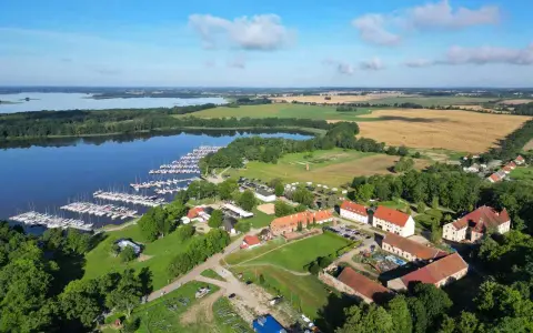Malowniczy widok na jezioro z przystanią pełną żaglówek, idealne miejsce na długi weekend czerwcowy. W oddali pola i zabudowania.