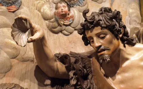 Detalle de escultura barroca: figura masculina con barba y cabello rizado sostiene una concha. Ángeles infantiles observan. Maestría de escultores del barroco.