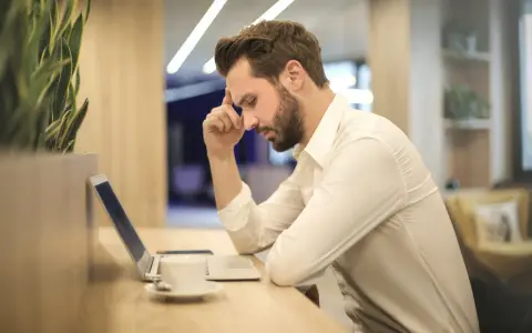 Mann mit Bart sitzt nach langer Krankheit wieder arbeiten ohne Wiedereingliederung am Laptop, die Stirn gerunzelt.
