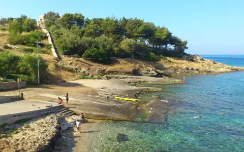 Scogliera e mare cristallino a San Gregorio Leuca. Persone si godono il sole e l'acqua, una scalinata sale verso la collina alberata.