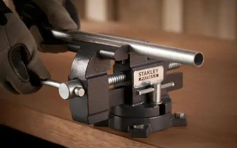 Manos enguantadas ajustan un tubo metálico en un tornillo de banco Stanley Fatmax, herramienta esencial para fijar piezas.