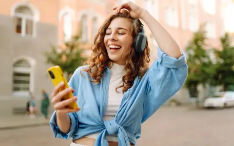Kobieta w słuchawkach i niebieskiej koszuli trzyma żółty telefon, słuchając muzyki.