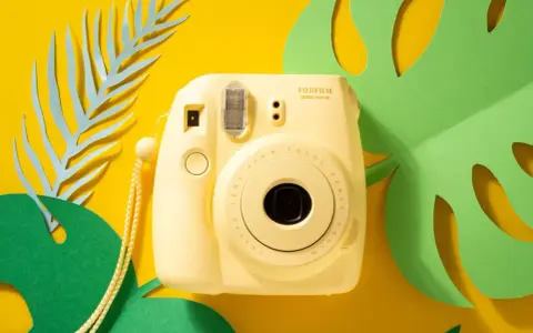 Żółty aparat natychmiastowy Fujifilm Instax mini 8 na tle tropikalnych liści.