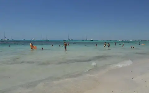 Słoneczny dzień na plaży Es Trenc. Ludzie kąpią się w turkusowej wodzie, a na horyzoncie widać żaglówki.