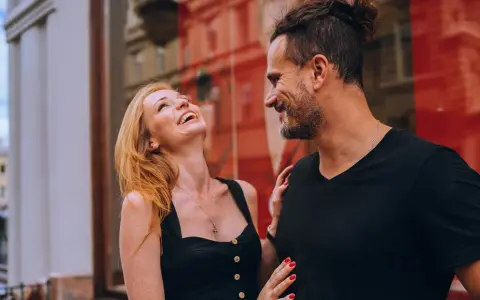Uśmiechnięta kobieta śmieje się głośno, podczas gdy zazdrosny facet milczy, patrząc na nią z czułością.