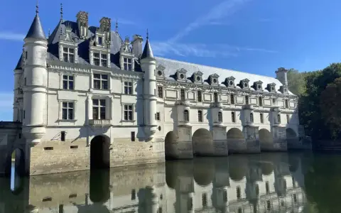 The stunning Chateau de Chenonceau, a white stone castle, gracefully spans the River Cher, its reflection shimmering on the water's surface under a clear blue sky.