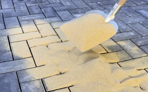 Schaufel mit Sand zum Terrassenplatten verfugen. Sand rieselt auf graue Pflastersteine.