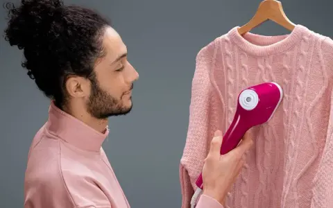 Man using a pink handheld steamer on a pink cable-knit sweater hanging on a wooden hanger.