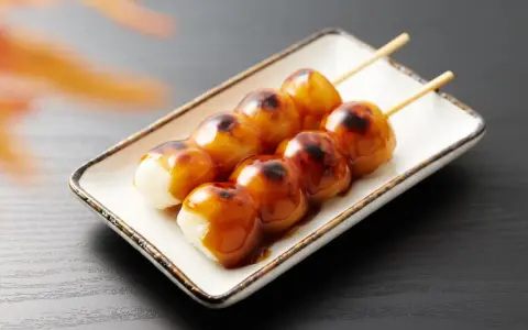 Brochettes de dango grillés, nappées de sauce sucrée, sur une assiette blanche.