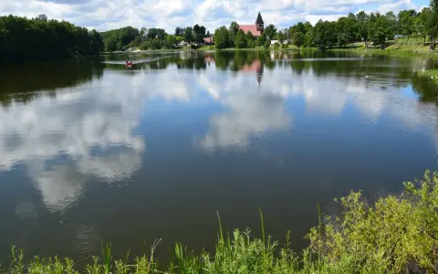 Spokojne jezioro na Kaszubach, odbijające chmury. W oddali widać łódkę i kościół. Idealne miejsce na wakacje.