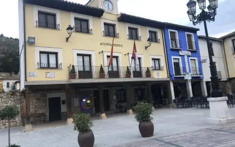 El Ayuntamiento de Tajuña, con su balcón y reloj, preside la plaza. Edificios de colores y farolas completan la escena.