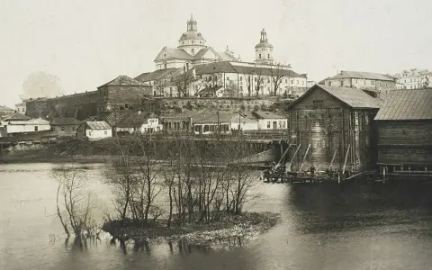 Widok na miasto z rzeką i kościołem. W oddali widać potężną budowlę sakralną z kopułami. Na pierwszym planie rzeka z wysepką i drzewami. Po prawej stronie drewniane budynki nad wodą. Ktoś pisze na Berdyczów.