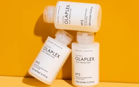 Drei Flaschen der OLAPLEX Serie, "Hair Perfector No.3", liegen übereinander auf einem orangen Hintergrund.