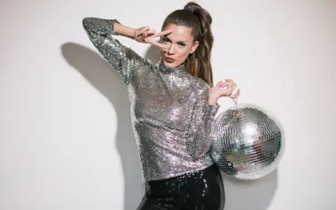 Kobieta w cekinowym topie i spodniach, z kulą dyskotekową w ręku, pozuje w stylu disco.