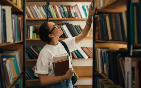 Młoda kobieta w okularach wybiera książkę z półki w bibliotece, trzymając inne w ręku.