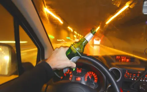 Ręka trzyma butelkę piwa za kierownicą. Po ilu można jechać autem po alkoholu? Nigdy!
