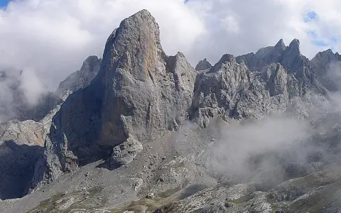 Majestatyczny **Naranjo de Bulnes** w Picos de Europa, otoczony mgłą i chmurami, z małym schroniskiem u podnóża.