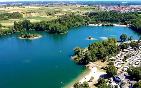 Der Mechtersheim See mit seiner türkisen Wasserfarbe, umgeben von grünen Bäumen und einem Campingplatz. Im Hintergrund erstrecken sich Felder und eine Stadt.