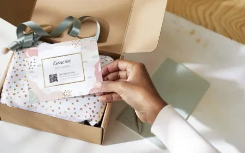 Tarjeta de agradecimiento tamaño postal con código QR, dentro de una caja de regalo abierta con papel de seda dorado.