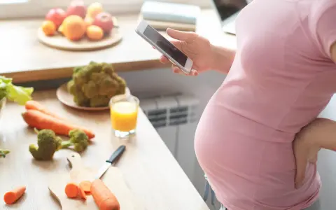 Ciężarna kobieta zmagająca się z brakiem apetytu w ciąży, szuka w telefonie przepisu na zdrowe jedzenie. Na blacie warzywa i sok.