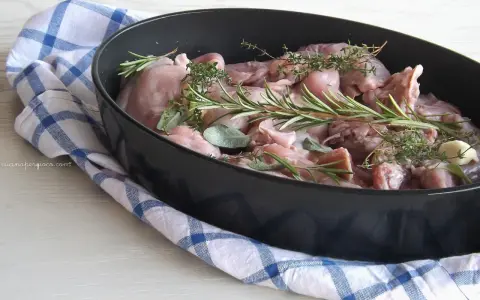 Pezzi di coniglio crudo pronti per essere cucinati al forno, profumati con rosmarino, salvia e aglio.