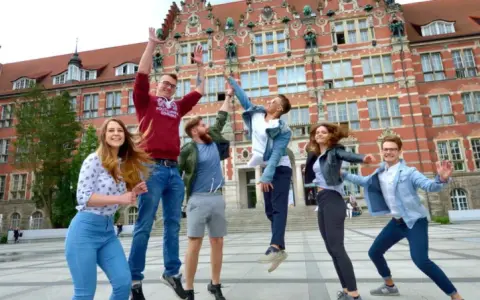 Radosna grupa studentów skacze przed budynkiem, gdzie można podjąć studia psychologiczne Gdańsk.