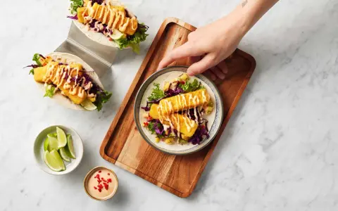Två läckra fish tacos med krispig fisk, rödkål, sallad och en krämig sås. En hand sträcker sig efter en tallrik med en taco.